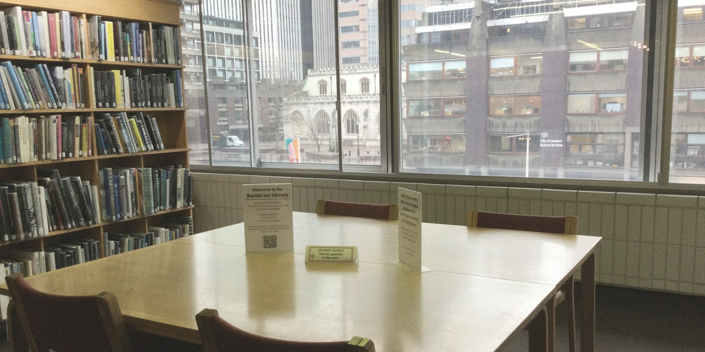 A quiet library reading room with shelves of books and a city view through the windows.